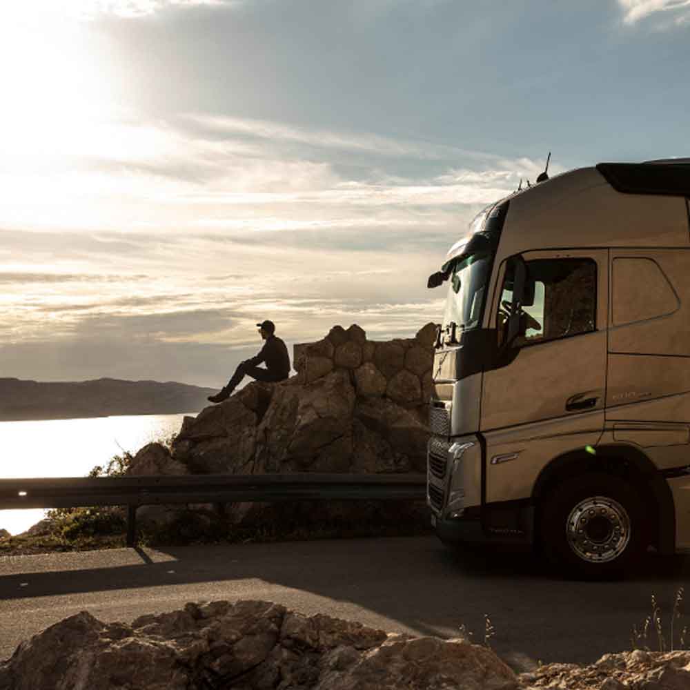 Vozač sjedi na stijeni i gleda more u zalazak sunca, a pored njega je parkiran kamion Volvo FH